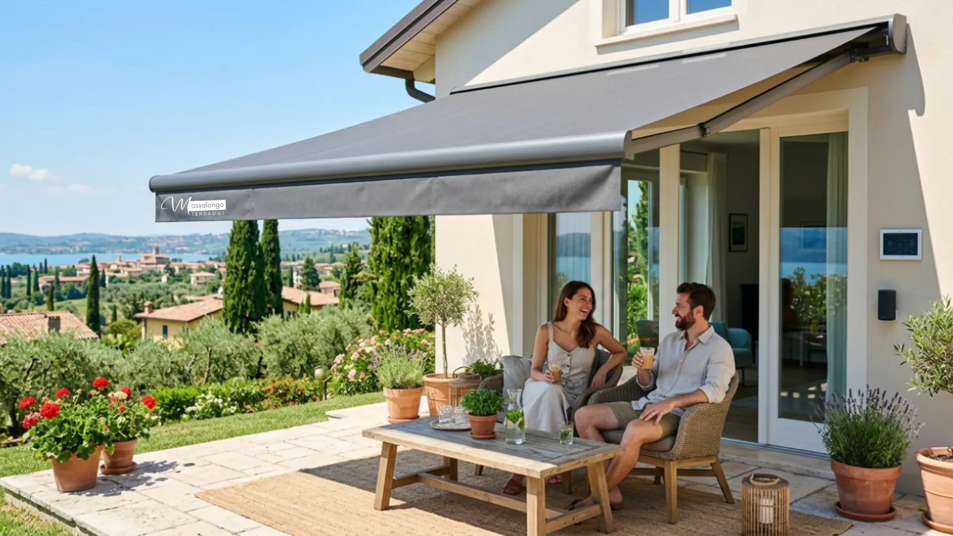 Una moderna terrazza pavimentata in pietra con due tende da sole a bracci Massalongo estese, che offrono ombra a una famiglia che cena a un tavolo di legno e a una coppia che si rilassa su sedie di vimini. La terrazza si affaccia su una vista panoramica sul Lago di Garda, con colline, cipressi e un villaggio italiano. La scena include vasi di terracotta con piante e un barbecue.